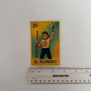 May include: A yellow and blue card with the number 25 and the text "EL PLOMERO" printed on it. The card features a cartoon illustration of a person wearing a blue shirt with a red lightning bolt symbol and blue jeans, holding a wrench and a blowtorch.