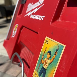 May include: A red Milwaukee Packout toolbox with a sticker of a Loteria card depicting a plumber holding a wrench and a pipe. The card is labeled "25 El Plomero".