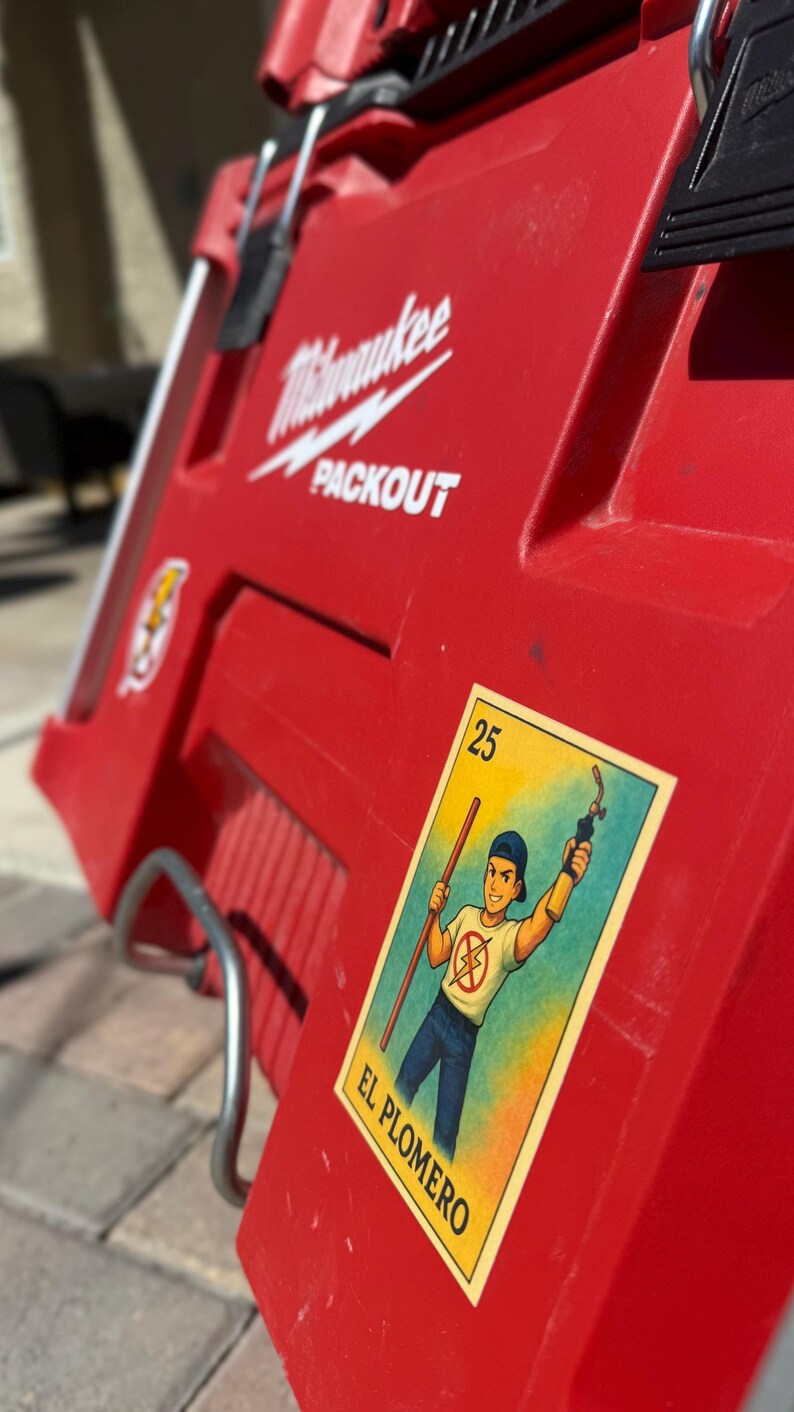 May include: A red Milwaukee Packout toolbox with a sticker of a Loteria card depicting a plumber holding a wrench and a pipe. The card is labeled "25 El Plomero".