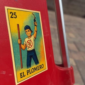 May include: A yellow and blue card with a cartoon illustration of a person holding a wrench and a pipe. The card has the number 25 and the text "El Plomero" on it.