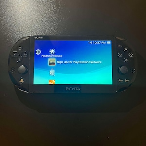 May include: A black Sony PlayStation Vita handheld gaming console with a bright screen displaying the PlayStation Network interface, including icons for the network, sign-up options, and the store, with the PS Vita logo printed at the bottom.