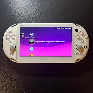 May include: A white Sony PlayStation Vita handheld gaming console with a bright screen displaying the PlayStation Network interface, including icons for the network and a sign-up prompt, with clear buttons and analog sticks on the sides.