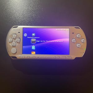 May include: A gold-colored Sony PlayStation Portable (PSP) handheld gaming console. The screen displays the PlayStation Network interface with the text "Sign Up for PlayStation Network." The console has a directional pad, buttons, and a power switch.