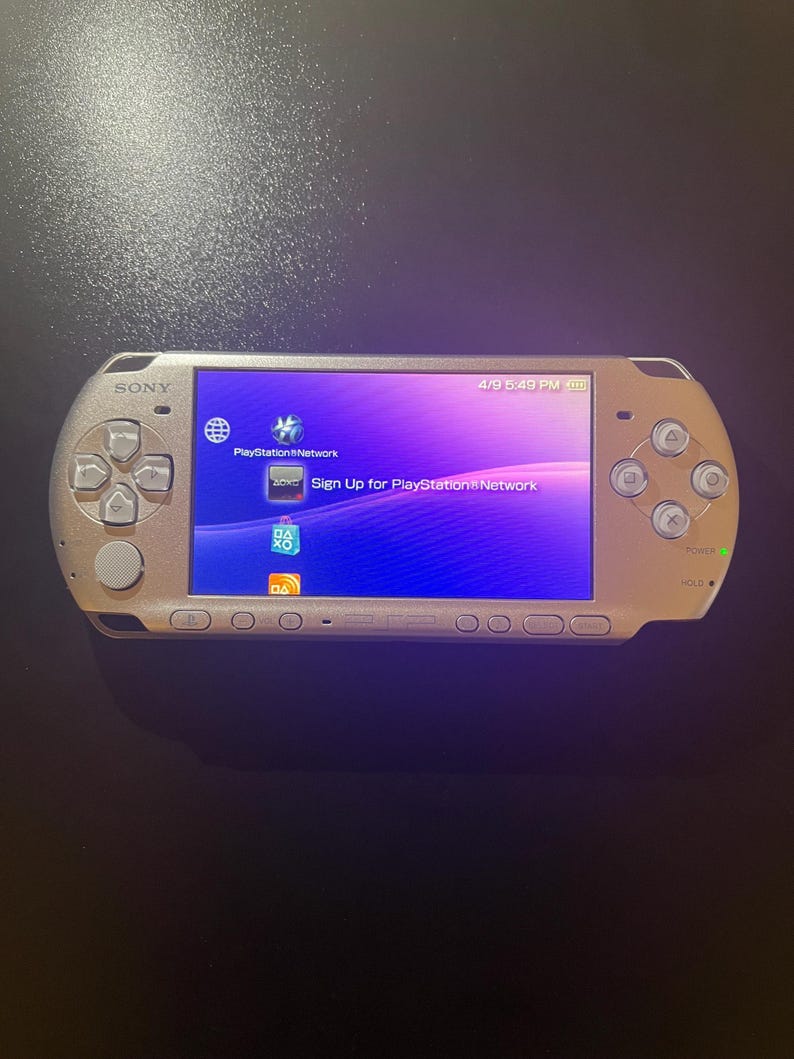 May include: A gold-colored Sony PlayStation Portable (PSP) handheld gaming console. The screen displays the PlayStation Network interface with the text "Sign Up for PlayStation Network." The console has a directional pad, buttons, and a power switch.