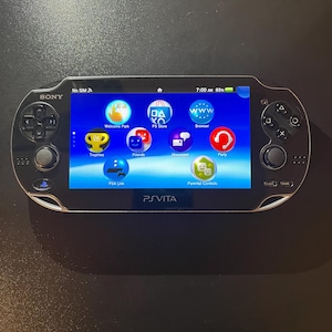 Op de afbeelding: Een zwarte Sony PlayStation Vita handheld spelcomputer met een glanzend scherm dat het hoofdmenu weergeeft. Het apparaat is voorzien van een richtingspad, twee analoge sticks en standaard actieknoppen. Het scherm toont iconen voor Welcome Park, PS Store, Browser, Trophies, Friends, Messages, Party, PS4 Link en Parental Controls.