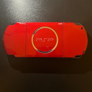 May include: A red Sony PSP handheld gaming console. The device features a circular emblem with the PSP logo in the center. The top of the console has the text "UMD". The Sony logo is visible on the left side.