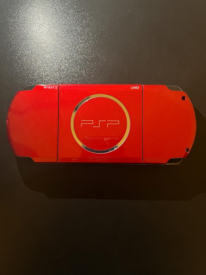 May include: A red Sony PSP handheld gaming console. The device features a circular emblem with the PSP logo in the center. The top of the console has the text "UMD". The Sony logo is visible on the left side.