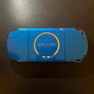 May include: A blue handheld gaming console with the text "PSP" in the center, surrounded by a gold circle. The console has the text "SONY" and "UMD" on the top. The device is on a dark surface.