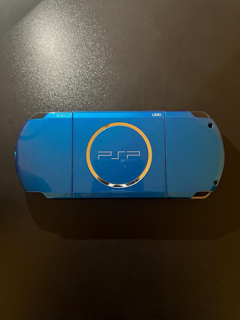May include: A blue handheld gaming console with the text "PSP" in the center, surrounded by a gold circle. The console has the text "SONY" and "UMD" on the top. The device is on a dark surface.
