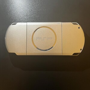 May include: A silver-colored PSP handheld gaming console. The device has a circular center with the PSP logo and a metallic finish. The console is rectangular with rounded edges and a smooth surface.