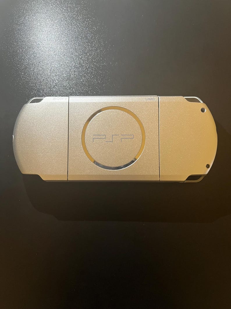 May include: A silver-colored PSP handheld gaming console. The device has a circular center with the PSP logo and a metallic finish. The console is rectangular with rounded edges and a smooth surface.
