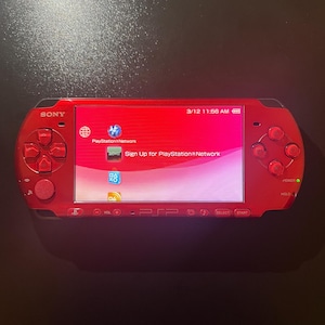 May include: A red Sony PSP handheld gaming console. The screen displays a pink and white interface with the text "PlayStation Network" and "Sign Up for PlayStation Network". The console has a directional pad, buttons, and a power button.