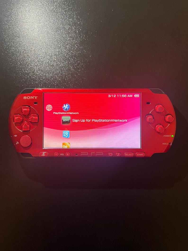 May include: A red Sony PSP handheld gaming console. The screen displays a pink and white interface with the text "PlayStation Network" and "Sign Up for PlayStation Network". The console has a directional pad, buttons, and a power button.