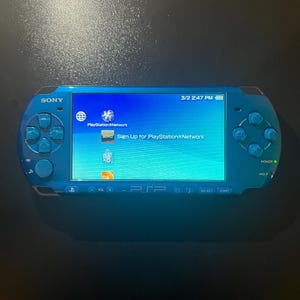 May include: A blue Sony PSP handheld gaming console. The screen displays the PlayStation Network sign-up page. The console has a directional pad, buttons, and a power switch. The date and time are visible on the screen.