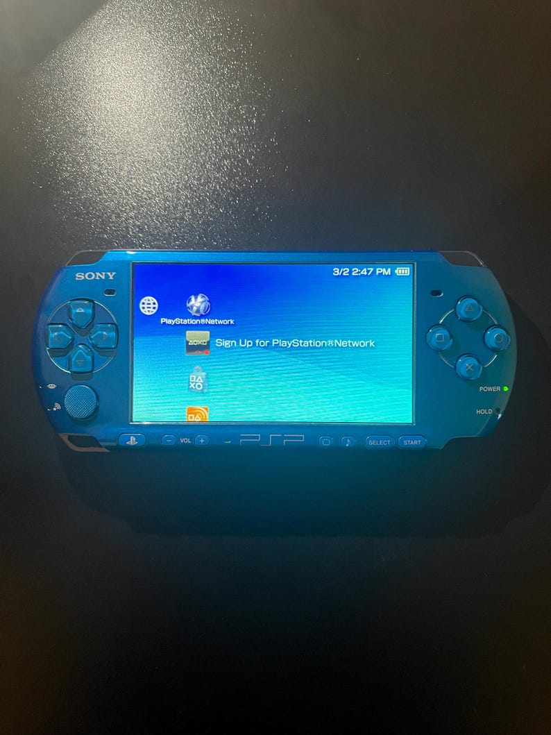 May include: A blue Sony PSP handheld gaming console. The screen displays the PlayStation Network sign-up page. The console has a directional pad, buttons, and a power switch. The date and time are visible on the screen.