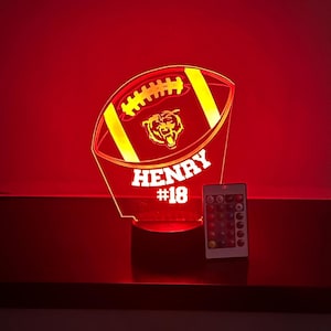 Puede incluir: Una lámpara roja iluminada con forma de balón de fútbol con el nombre "HENRY" y el número "18" en blanco. La lámpara presenta un logotipo de oso y detalles amarillos. Un mando a distancia con opciones de color está cerca.