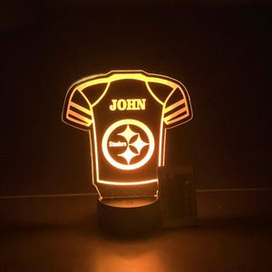 May include: A yellow and black illuminated night light shaped like a football jersey with the name "JOHN" and the Pittsburgh Steelers logo. The light is on a black base with a switch.