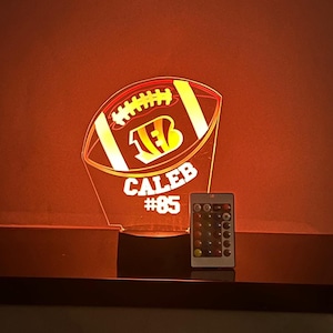 May include: A glowing football-shaped night light with the name "CALEB" and the number "85" displayed. The light features the team logo and comes with a remote control. The light is on a dark surface against an orange background.
