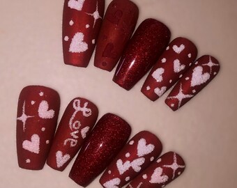 May include: Red and white press-on nails with a Valentine's Day theme. The nails feature white hearts, glitter, and the word "Love" written on one nail.