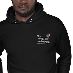 Peut inclure: Sweat à capuche noir avec le texte "You have to gain people's trust before you can manipulate them" et des graphiques d'yeux brodés. Le sweat à capuche a une capuche à cordon et une poche avant.