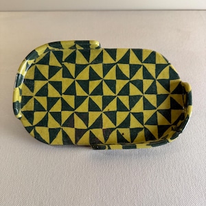 May include: A ceramic tray with a geometric pattern of yellow and dark green triangles. The tray has a rectangular shape with rounded edges and a small handle on one side. The design is repeated across the surface.