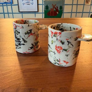 May include: Two ceramic mugs with a speckled design in red, black, and green on a cream background. The mugs have a unique handle design and sit on a wooden table. The mugs are part of a kitchenware set.