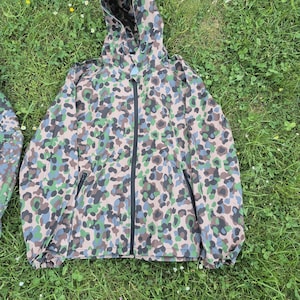 Könnte beinhalten: Eine Tarnjacke mit Kapuze und durchgehendem Reißverschluss. Die Jacke hat ein Muster aus grünen, blauen und braunen Formen auf beigem Hintergrund. Sie hat zwei Vordertaschen und liegt auf grünem Gras.