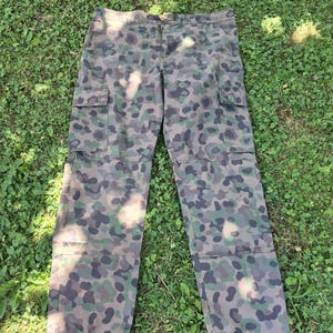 Può includere: Pantaloni cargo con motivo mimetico nei toni del marrone, verde scuro e nero. I pantaloni presentano una vita con passanti per cintura, una chiusura frontale con cerniera e due ampie tasche cargo laterali con patta posizionate sulle cosce.