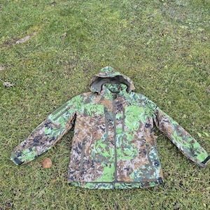 Può includere: Una giacca mimetica verde e marrone con cappuccio e chiusura a zip. La giacca ha più tasche e un cappuccio con coulisse.