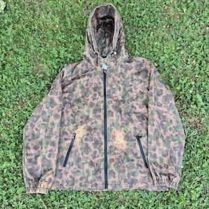 Könnte beinhalten: Eine Kapuzenjacke mit Camouflage-Muster in Braun-, Grün- und Beigetönen. Die Jacke verfügt über einen durchgehenden schwarzen Frontreißverschluss, zwei seitliche Reißverschlusstaschen und elastische Bündchen. Die Kapuze hat einen Kordelzug.