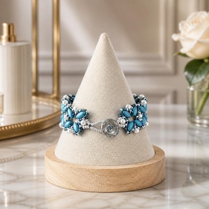 May include: A beaded bracelet featuring blue marquise-shaped beads and small white pearls arranged in a star-like pattern. The bracelet is secured with a circular silver-toned button clasp and is displayed on a beige cone-shaped jewelry stand.