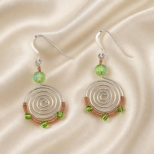 Può includere: Un paio di orecchini in filo metallico color argento con design a spirale. Ogni orecchino include una perla verde chiaro nella parte superiore e tre perline verdi più piccole fissate alla curva inferiore con filo color rame e ganci argentati.
