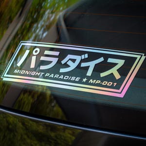 May include: A rectangular holographic decal applied to a car window. The sticker features the Japanese text "パラダイス" in large characters, with the English text "MIDNIGHT PARADISE ★ MP-001" underneath, all enclosed within a double-lined rectangular border.