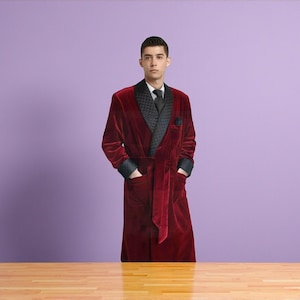 May include: A burgundy velvet robe with black satin lapels and cuffs. The robe has a matching belt and pockets. The person is wearing a black tie and a white shirt. The background is a solid purple color.
