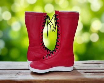 May include: A pair of red, high-top boots with black laces. The boots have a white sole and are made of a smooth material. The boots are displayed on a wooden surface against a blurred green background.