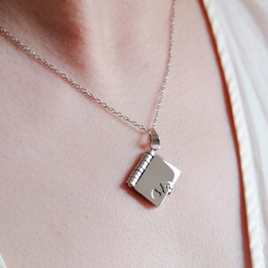 Puede incluir: Collar plateado con un pequeño colgante cuadrado. El colgante tiene forma de libro en miniatura con encuadernación en espiral y un pequeño cierre. El colgante tiene un pequeño corazón y el número 36 grabado.