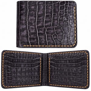 May include: A black bifold wallet featuring a textured crocodile skin pattern and contrasting tan stitching along the edges. The wallet is shown in both a closed position and an open position, revealing multiple interior card slots on each side.