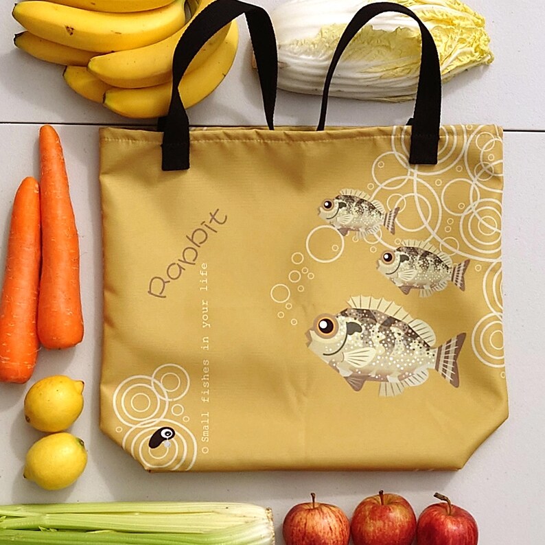 May include: A mustard-colored tote bag with black handles, featuring a fish design and the word "Rabbit." The bag is surrounded by fresh produce, including carrots, lemons, apples, bananas, and celery.
