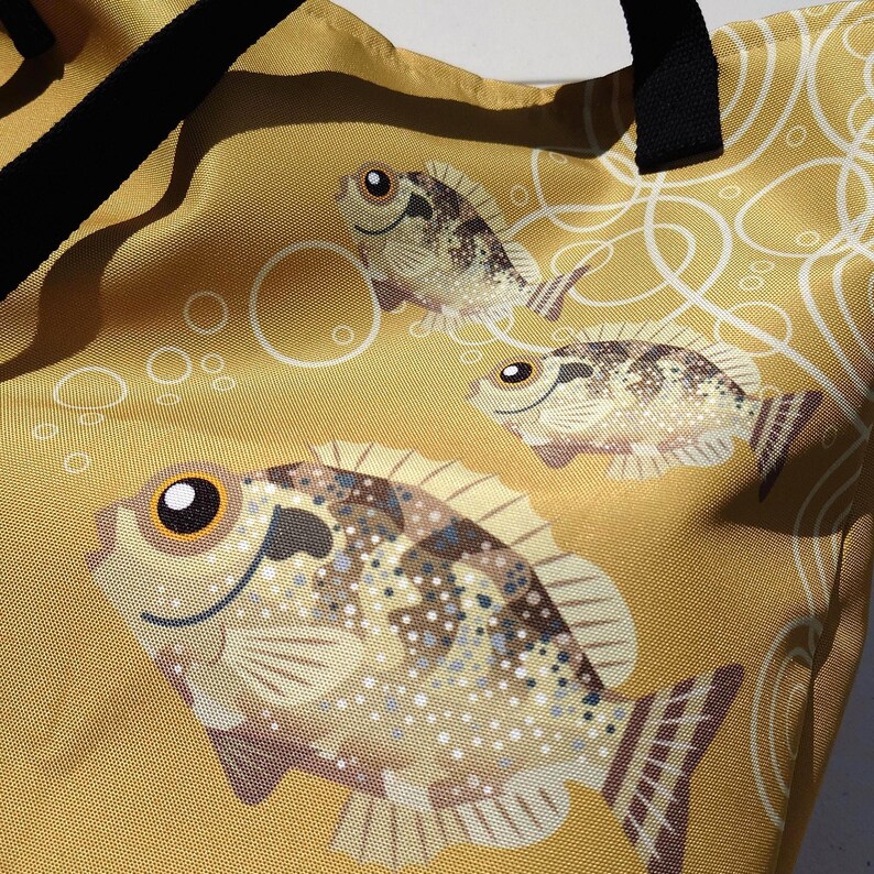 May include: A mustard yellow tote bag featuring a repeating pattern of cartoon fish and bubbles. The fish have brown and tan speckled bodies with large, expressive eyes. Black handles are visible at the top of the bag.