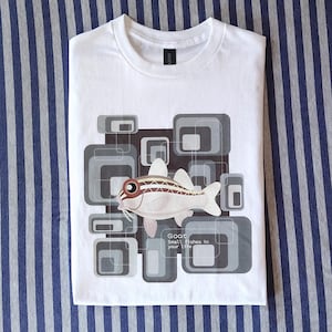 Op de afbeelding: Een wit t-shirt met een afbeelding van een vis met bruine en witte strepen. De achtergrond bestaat uit overlappende grijze en donkergrijze afgeronde rechthoeken. De tekst luidt: Goat, Small fishes in your life. Het ontwerp is gecentreerd.