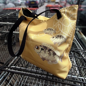 May include: A mustard yellow tote bag featuring a repeating pattern of fish and bubbles. The bag has black handles and a black interior. The fish are brown and white with large eyes. The bag is sitting in a shopping cart.
