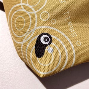 May include: Close-up of a gold-colored fabric with a repeating pattern of white circles and a cartoon-style black and white character. The character has a teardrop detail. The word "Small" is printed on the fabric.