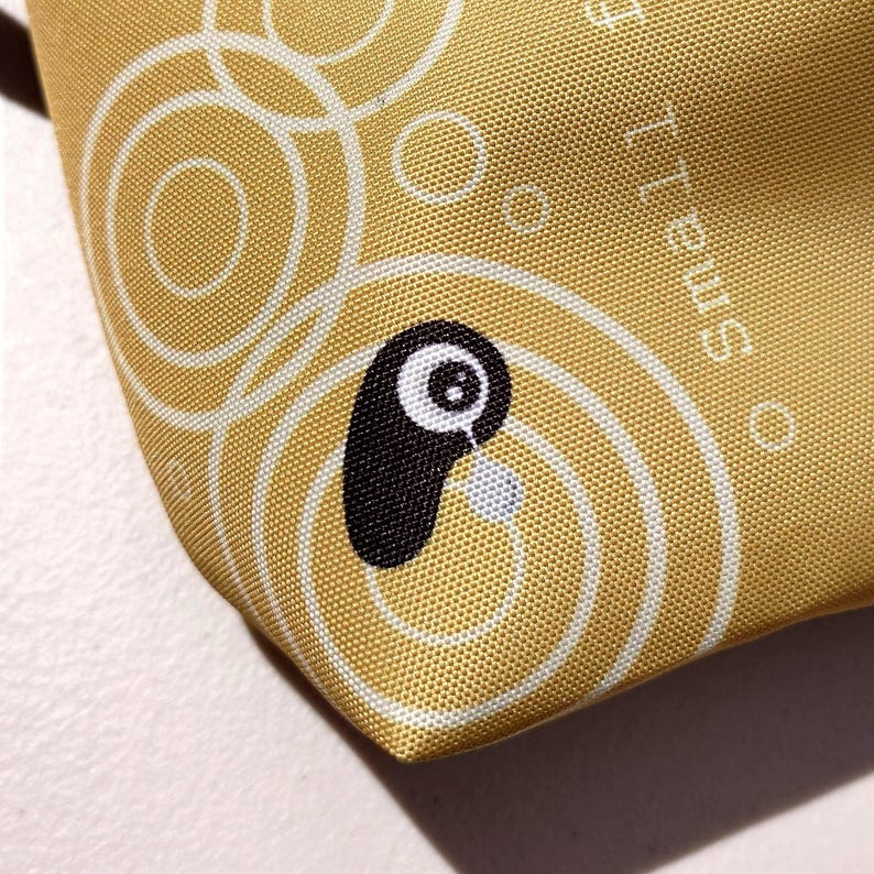 May include: Close-up of a gold-colored fabric with a repeating pattern of white circles and a cartoon-style black and white character. The character has a teardrop detail. The word "Small" is printed on the fabric.