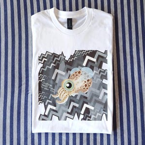 May include: A white short-sleeved t-shirt featuring a graphic of a tan and brown spotted cuttlefish centered over a geometric pattern of gray, black, and white zigzags. The text "Cuttle Small fishes in your life" is printed in white on the left.