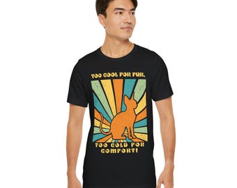 May include: A black t-shirt featuring a retro-style graphic of a hairless cat. The graphic has a rainbow background with the text "Too Cool For Fur, Too Cold For Comfort!"