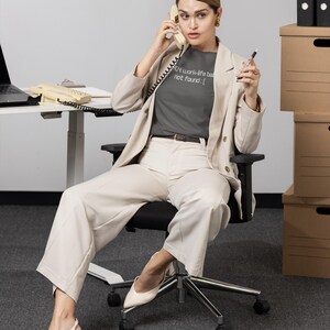 May include: A person wearing a gray t-shirt with the text "404 work-life balance not found:[" paired with a beige blazer and pants. The person is seated in a black office chair, holding a phone and pen.