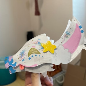 May include: A handmade, whimsical paper mask in the shape of a stylized animal head. The mask is primarily white with colorful accents, including blue, pink, gold, and a yellow star. The ears have pink interiors and blue accents.