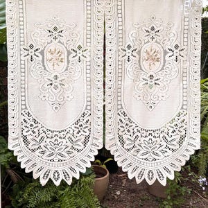 Könnte beinhalten: Zwei cremeweiße Spitzengardinen mit aufwendiger Blumenstickerei in der Mitte, kunstvollen Schnörkelmustern und einem gewellten unteren Rand. Der Stoff hat ein strukturiertes Netzdesign mit einer dekorativen Bordüre an den Seiten.