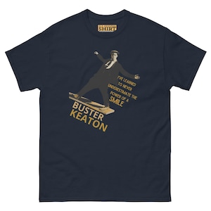 Puede incluir: Una camiseta azul marino con un gráfico de Buster Keaton en un monopatín. El texto "He aprendido a nunca subestimar el poder de una sonrisa" está impreso en dorado en la camiseta. El texto "Buster Keaton" también está impreso en dorado en la camiseta.
