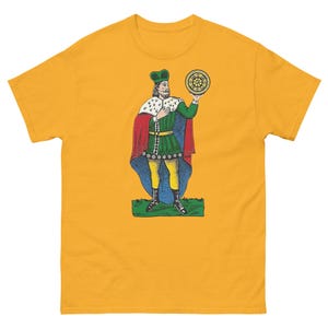 Puede incluir: Una camiseta amarilla con una ilustración colorida de un rey sosteniendo un orbe dorado. El rey lleva una corona verde y dorada, una túnica roja y verde y pantalones amarillos. El orbe está decorado con un patrón de estrella.